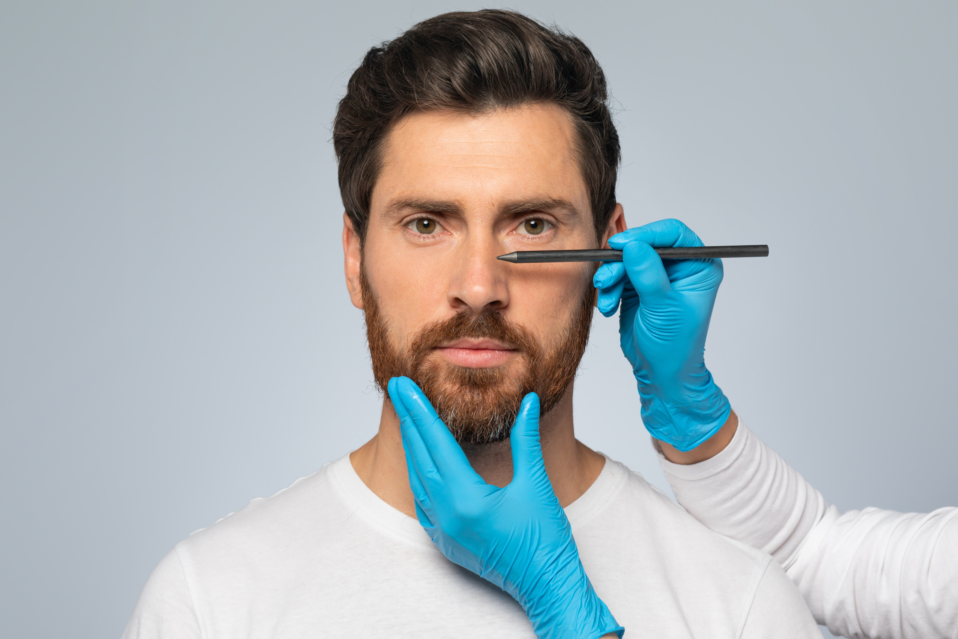 Doctor making marks on male patient's face, middle aged man on consultation at surgeon, standing on grey background